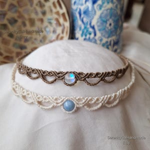 May include: Two macrame chokers featuring intricate knotted patterns. The top choker is brown with a round, iridescent moonstone. The bottom choker is cream-colored with a round, light blue stone. Both necklaces have a delicate, scalloped edge.