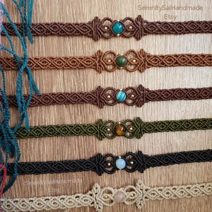 May include: Macrame chokers in various colors, including brown, green, and black, each featuring a central gemstone. The chokers have intricate knotting patterns and are adorned with small gold-colored beads. The Etsy shop name "SerenitySalHandmade" is visible.