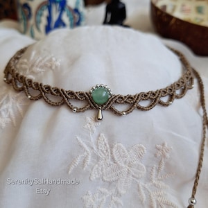 May include: A macrame choker necklace crafted from brown cord featuring a central round green gemstone, small silver-toned metallic beads, and a single silver-toned teardrop-shaped pendant hanging from the center of the woven pattern.