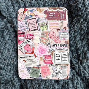 May include: A decorative case covered in stickers with a book theme. The stickers include phrases like "Cozy Girl Book Club," "Booktok Made Me Read It," and "He's a 10 but..." The case is set against a textured, blue-gray background.