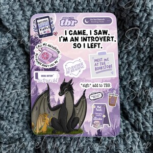May include: A rectangular sticker with a purple background and various illustrations and text. The sticker features a black dragon, a yellow dragon, and phrases like "I came, I saw, I'm an introvert, so I left." and "I'd rather be reading."