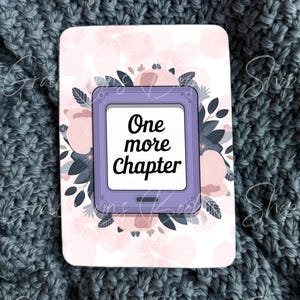 May include: A rectangular sticker featuring a purple e-reader device in the center with the text One more Chapter displayed on its screen. The device is surrounded by a decorative pattern of soft pink flowers and dark blue-grey leaves on a white background.