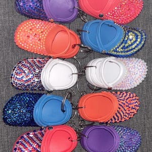 Puede incluir: Una colección de mini llaveros de zuecos enjoyados en varios colores. Los llaveros presentan una gama de colores, incluyendo rosa, morado, azul, naranja y blanco, con brillantes adornos de pedrería. Cada llavero tiene un anillo de metal.