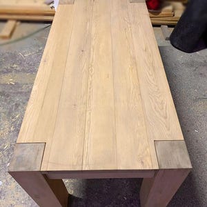 Puede incluir: Una mesa de madera clara con una tapa rectangular y cuatro patas. La tapa de la mesa está hecha de tablones de madera que se unen. La mesa está sin terminar y tiene un color de madera natural.