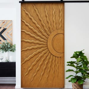 Könnte beinhalten: Eine große Holzschiebetür mit einem eingearbeiteten Sonnenmuster. Die Tür hat einen warmen, natürlichen Holzton und hängt an einer schwarzen Metallschiene. Ein rosa-rot gestreifter Teppich liegt vor der Tür. Die Tür ist etwa 213 cm hoch.