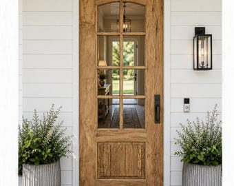 May include: A wooden front door with an arched top and six glass panes, featuring a rectangular panel at the bottom. The door is set within a white horizontal siding wall, flanked by two large, ribbed gray planters containing green plants.