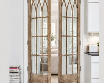 May include: A pair of light brown wooden double doors featuring glass panels with a grid pattern and arched tops. The doors are set within a white door frame, revealing a glimpse of a bright interior space with white walls and flooring.