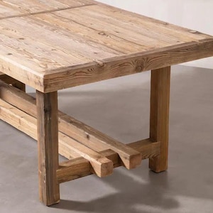 Pode incluir: Mesa de madeira rústica com tampo retangular e pernas robustas. A mesa é construída em madeira clara, mostrando grãos e nós visíveis. O design apresenta uma base em estilo cavalete, adicionando à sua estética de fazenda.