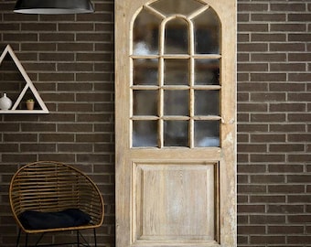 May include: A light-toned wooden sliding barn door featuring an arched top with twelve glass panes and a solid rectangular lower panel. The door is mounted on a black metal rail system against a dark brown brick wall with a visible wood grain finish.
