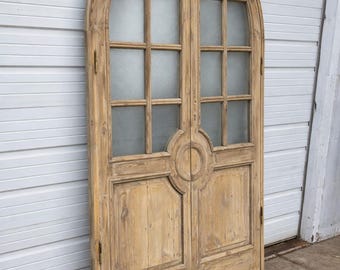 Può includere: Un paio di antiche porte in legno con la parte superiore ad arco. Le porte sono divise in pannelli con vetri smerigliati. Il legno ha una finitura marrone chiaro usurata. Le porte sono appoggiate a un muro bianco.
