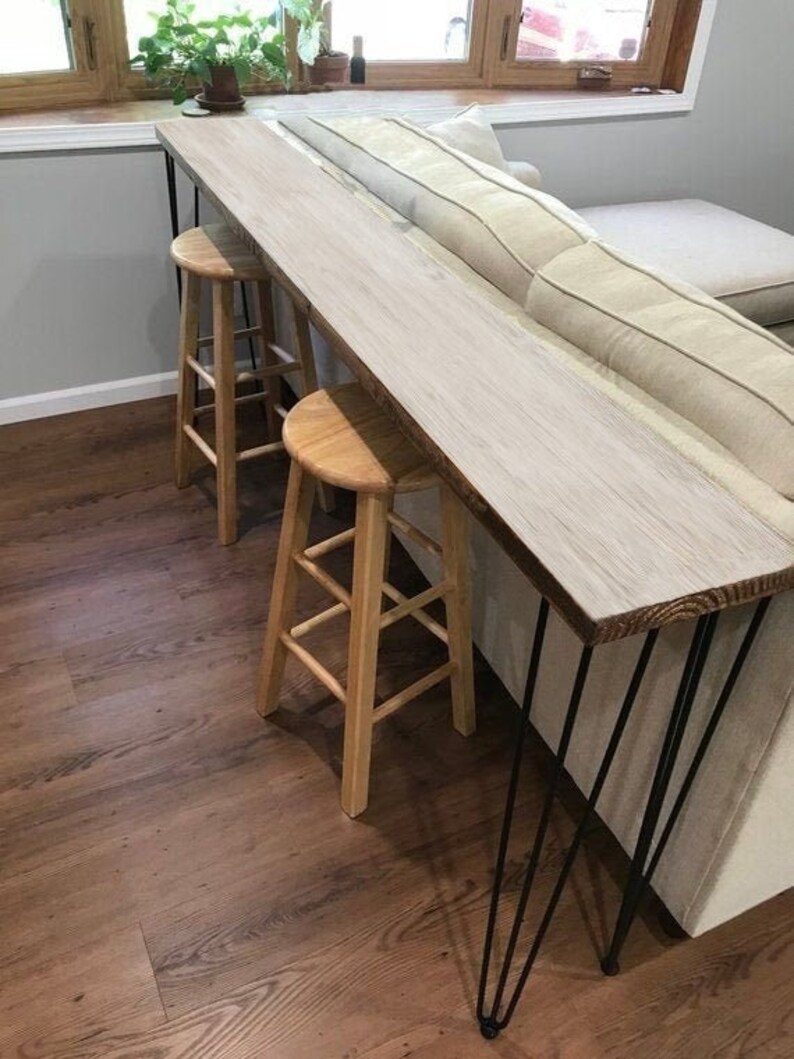 May include: A whitewashed wood console table with black hairpin legs. The table is positioned next to a white couch and has two wooden stools placed underneath.