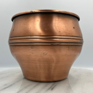 Könnte beinhalten: Ein Kupfertopf mit rundem Körper und leicht ausgestelltem Rand. Der Topf hat drei horizontale Bänder um die Mitte.