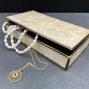 Può includere: Un portagioie rettangolare con coperchio e lati color crema effetto marmo, rifinito con bordi dorati. All'interno, sono visibili una collana di perle bianche e una collana con pendente dorato. La scatola è parzialmente aperta.