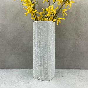 May include: White ceramic vase with a textured surface. The vase is tall and narrow, with a rectangular shape. It is filled with yellow flowers.