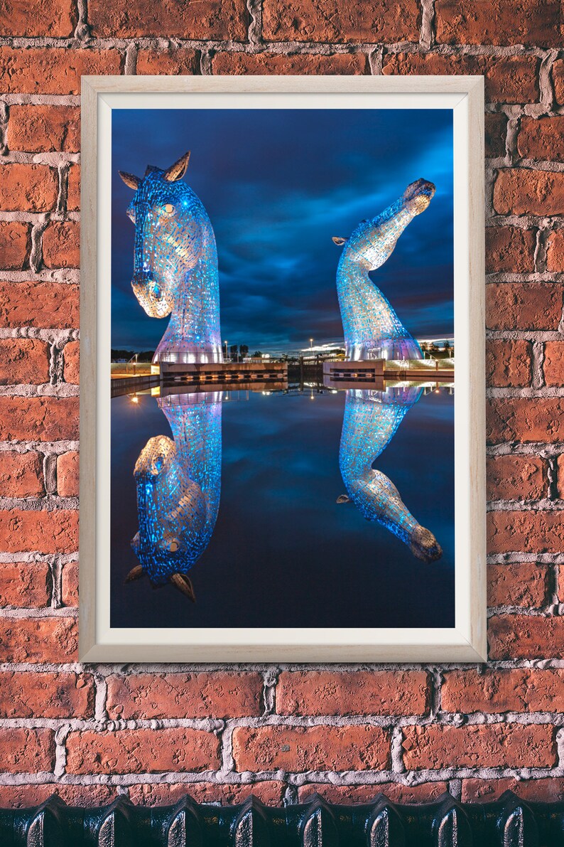 May include: Two blue, metallic horse sculptures stand in a body of water, reflecting the night sky. The sculptures are illuminated with blue lights, creating a vibrant and modern art piece.