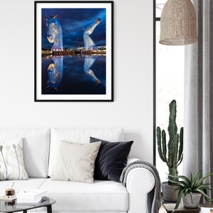 May include: A framed photograph of a large metal sculpture of two horses' heads, illuminated with blue lights, reflected in a still body of water at night.