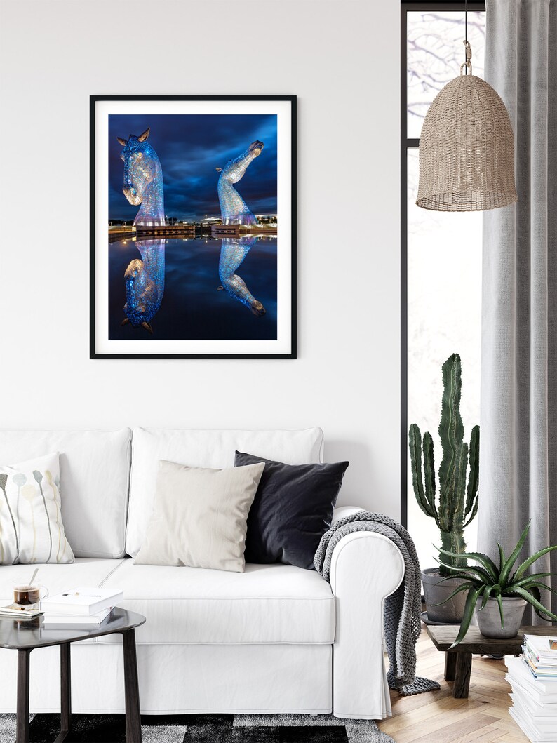 May include: A framed photograph of a large metal sculpture of two horses' heads, illuminated with blue lights, reflected in a still body of water at night.