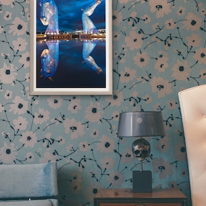 May include: A framed photograph of a blue sculpture of two horses reflected in a body of water. The sculpture is lit up at night. The photo is hanging on a wall with a floral pattern.