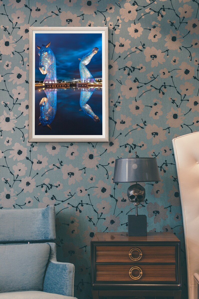 May include: A framed photograph of a blue sculpture of two horses reflected in a body of water. The sculpture is lit up at night. The photo is hanging on a wall with a floral pattern.