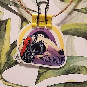 May include: A sticker featuring a red and black frog climbing on a purple skull, set against a rainbow background. The sticker is held by a yellow binder clip and twine, with a leafy background.