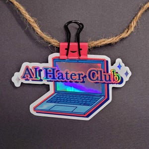 May include: A sticker with a laptop design and the text "AI Hater Club" in a holographic style. The sticker is held by a pink binder clip, with a brown twine string attached.