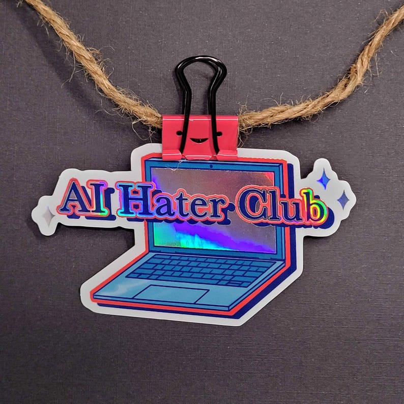 May include: A sticker with a laptop design and the text "AI Hater Club" in a holographic style. The sticker is held by a pink binder clip, with a brown twine string attached.