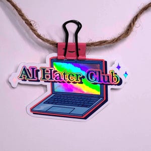 May include: A holographic sticker with a laptop design and the text "AI Hater Club." It's held by a pink binder clip and suspended by a brown string against a white backdrop. The laptop screen displays a rainbow gradient.