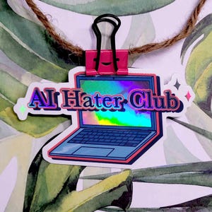 May include: A sticker with a laptop design and the text "AI Hater Club" in a holographic font. It's held by a pink binder clip, with a natural twine string. The background features a watercolour leaf design.
