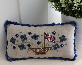 May include: A blue and white embroidered pillow with a floral design and a small flag in the center. The pillow has a blue velvet trim.