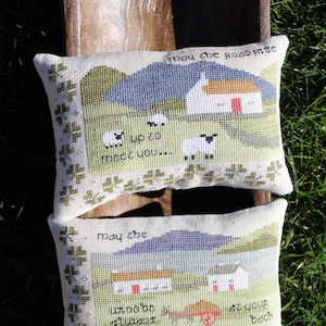 Puede incluir: Dos fundas de coj&iacute;n bordadas con bendiciones irlandesas. La almohada superior dice "Que el camino se eleve para encontrarte..." y presenta un paisaje verde con una casa blanca y ovejas. La almohada inferior dice "Que el viento siempre est&eacute; a tu espalda" y presenta un paisaje similar con una casa blanca y un carro rojo.