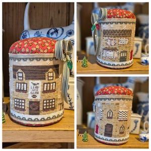 May include: A cylindrical pincushion with a red patterned top and a cross-stitched exterior depicting a building. The building has windows, doors, and text. A striped ribbon and a green gem adorn the side. The pincushion sits on a wooden shelf.