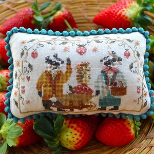 Könnte beinhalten: Ein rechteckiges, besticktes Kissen mit türkisfarbenem Bommelbesatz, das Waschbären und Erdbeeren zeigt. Das Kissen ist von frischen Erdbeeren in einem Weidenkorb umgeben.