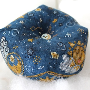 May include: A dark blue pincushion with a celestial design. The fabric is embroidered with gold, white, and silver stars, constellations, and a sun and moon motif. A small button is centered on the top.