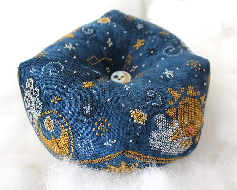 May include: A dark blue pincushion with a celestial design. The fabric is embroidered with gold, white, and silver stars, constellations, and a sun and moon motif. A small button is centered on the top.
