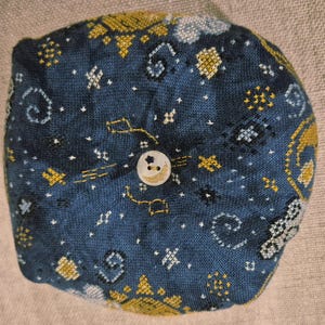 May include: A round, dark blue fabric item with embroidered celestial patterns in gold and white. A small, round button with a star and crescent moon design is centered on the fabric.