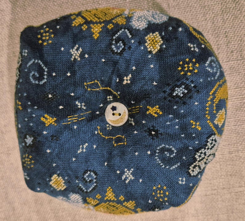May include: A round, dark blue fabric item with embroidered celestial patterns in gold and white. A small, round button with a star and crescent moon design is centered on the fabric.