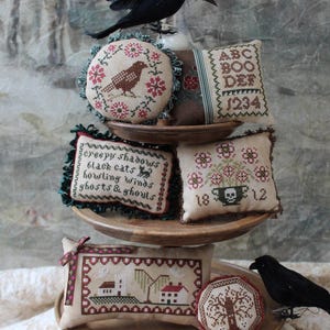 May include: A tiered wooden stand displays a collection of five embroidered pillows with Halloween themes. The pillows feature images of crows, skulls, and text like "Creepy Shadows, Black Cats, Howling Winds, Ghosts & Ghouls" and "ABC BOO DEF 1234".