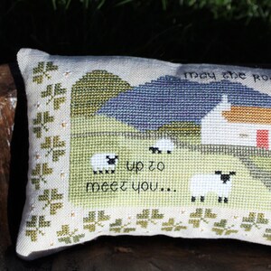 Puede incluir: Una almohada blanca con un dise&ntilde;o de punto de cruz de una caba&ntilde;a, colinas y ovejas. El texto "May the road rise up to meet you..." est&aacute; bordado en la almohada.