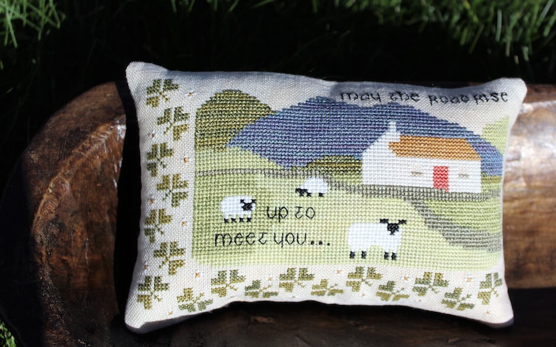 Puede incluir: Una almohada blanca con un dise&ntilde;o de punto de cruz de una caba&ntilde;a, colinas y ovejas. El texto "May the road rise up to meet you..." est&aacute; bordado en la almohada.