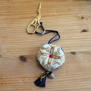Op de afbeelding: Een witte en gouden naaldhouder met een rode knop in het midden. De naaldhouder is bevestigd aan een blauw koord met een gouden schaar en een gouden vingerhoed.