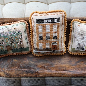Może przedstawiać: Trzy dekoracyjne, prostokątne poduszki z haftowanymi wzorami domów. Każda poduszka ma beżowe tło, szczegółowe przeszycia i obszycie z koralików. Poduszki są prezentowane w drewnianej misie.