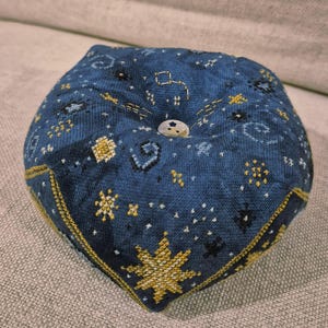 May include: A blue pincushion with a celestial design. The fabric is embroidered with gold and white stars, constellations, and other celestial elements. A white button is in the center. The pincushion is approximately 4 inches wide.