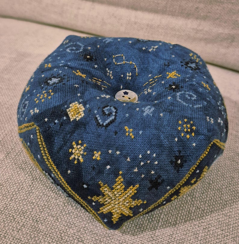 May include: A blue pincushion with a celestial design. The fabric is embroidered with gold and white stars, constellations, and other celestial elements. A white button is in the center. The pincushion is approximately 4 inches wide.
