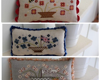 Puede incluir: Tres fundas de cojín bordadas con diferentes diseños florales. La almohada superior tiene un borde festoneado rojo y blanco y presenta una abeja y flores en una maceta marrón. La almohada del medio tiene un borde de terciopelo azul y presenta flores azules en una maceta marrón. La almohada inferior tiene un borde festoneado de color melocotón y presenta flores blancas en una maceta a cuadros blancos y negros con un letrero que dice "Melocotones frescos".