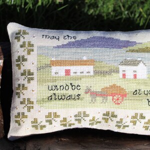 Puede incluir: Una almohada blanca con un borde de tr&eacute;bol verde y un dise&ntilde;o de punto de cruz de una escena campestre con dos casas, un carro y el texto "Que el viento siempre est&eacute; a tu espalda".