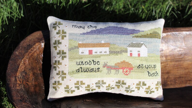 Puede incluir: Una almohada blanca con un borde de tr&eacute;bol verde y un dise&ntilde;o de punto de cruz de una escena campestre con dos casas, un carro y el texto "Que el viento siempre est&eacute; a tu espalda".