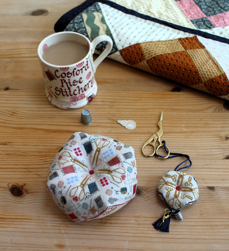 Op de afbeelding: Een naaldkussen van naaldkunst met een ontwerp van scharen, knopen en andere naai-accessoires. Het naaldkussen ligt op een houten tafel met een kopje thee, een vingerhoed en een schaar.