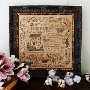 May include: A framed cross-stitch sampler with a dark brown ornate frame. The sampler features a house, flowers, and text that reads "Magnolia blooms and cotton fields...the heart of home you'll find." The year 2025 is also visible.