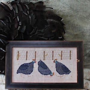 May include: A framed cross-stitch embroidery featuring three blue guinea fowl standing behind a wire fence. The birds are detailed with black and white speckles and have yellow beaks and wattles.