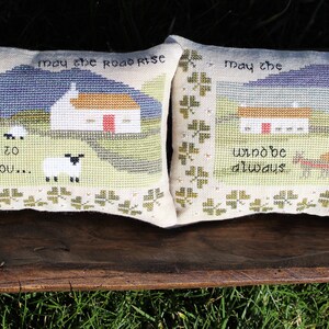 Puede incluir: Dos fundas de coj&iacute;n bordadas con una escena tradicional del campo irland&eacute;s. Las almohadas presentan una caba&ntilde;a blanca, colinas verdes y ovejas. El texto en las almohadas dice "Que el camino se eleve para encontrarte..." y "Que el viento est&eacute; siempre a tu espalda."